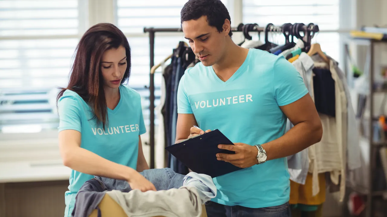 Pessoas voluntárias organizando roupas em um espaço de doações, usando camisetas azuis com a palavra “Volunteer”.
